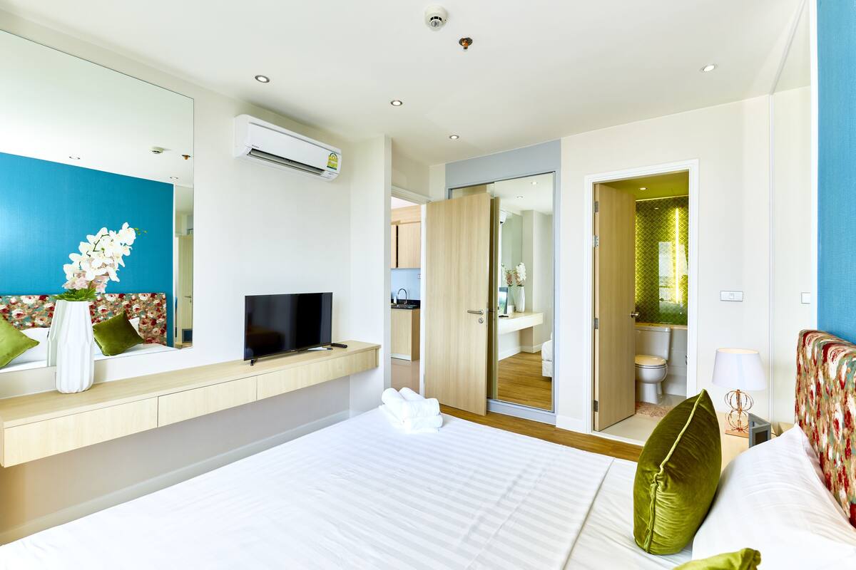 Ein modernes Hotelzimmer mit Bett, Fernseher, einem durchgehenden Türrahmen sichtbarem Badezimmer und einem Spiegel an der Wand.