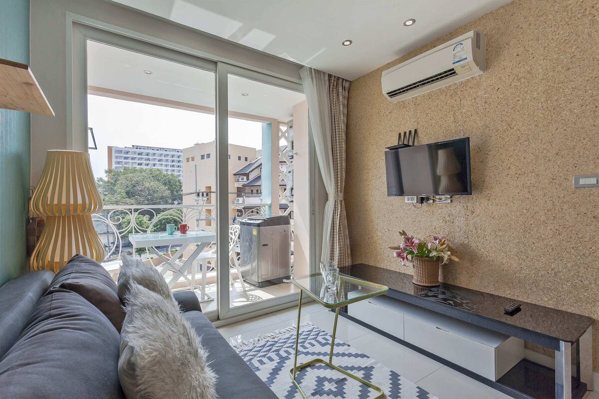 Ein modernes Wohnzimmer mit Sofa, Glastisch, Flachbildfernseher und einem Balkon mit Gartenmöbeln.