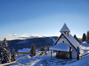 Un refugio de montaña cubierto de nieve, con estructura de madera, un porche techado y vistas a árboles y montañas nevadas.
