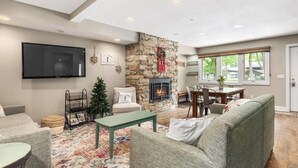 A spacious living room with a stone fireplace, a flat-screen TV mounted on the wall, and a cozy seating area with a green sofa and a patterned rug. - Boyne Mountain, Base of Mtn, Sleeps 13 (Boyne Falls)