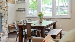 A dining area with a stone wall, a wooden table, metal chairs, and a view of a residential neighborhood through large windows. - Boyne Mountain, Base of Mtn, Sleeps 13 (Boyne Falls)