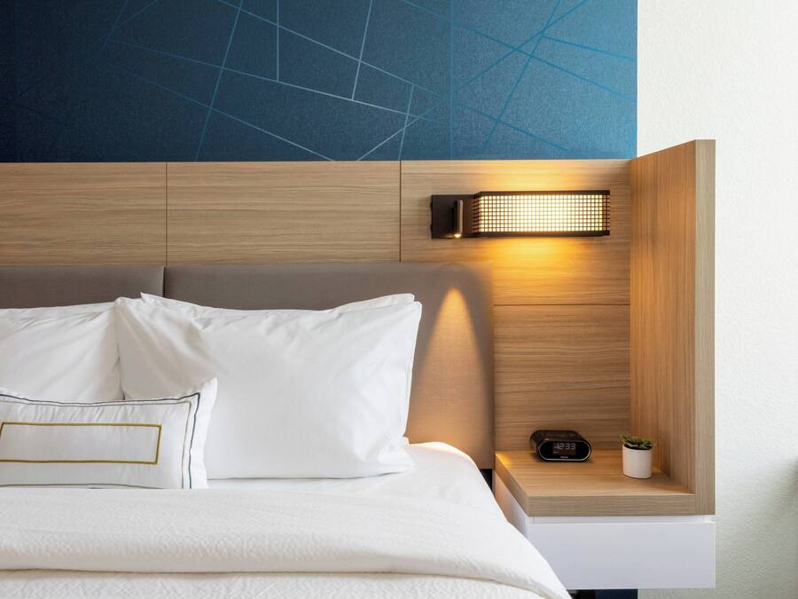 A neatly made bed with white linens and pillows, a bedside table with a clock and a small plant, and a wall-mounted light fixture.