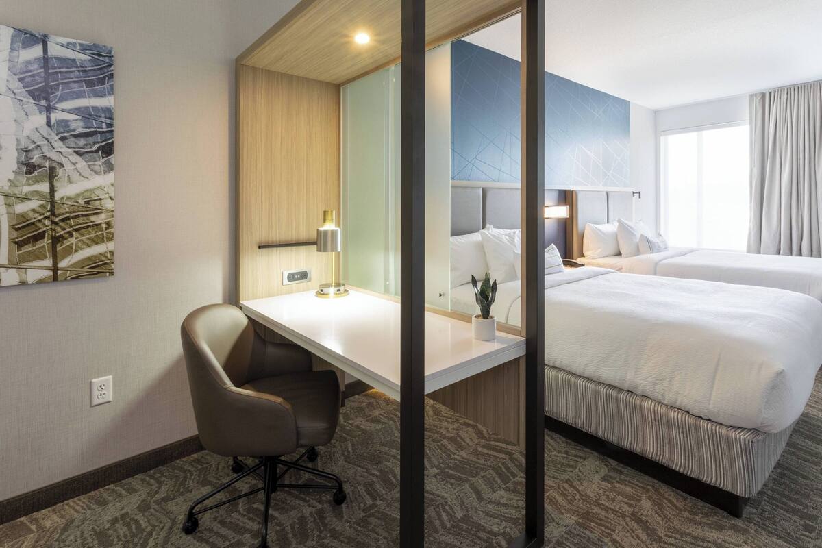 A hotel room with a desk, two beds, a chair, a lamp, and a large window.