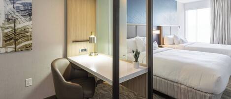 A hotel room with a desk, two beds, a chair, a lamp, and a large window.