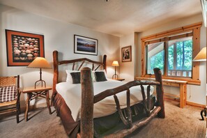 A rustic wooden bed with a view of trees through a window, a checkered chair, and framed pictures on the walls.