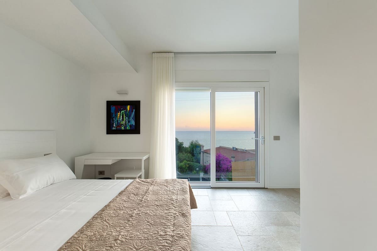 A modern bedroom with a bed, a desk, and a large sliding glass door leading to a balcony with a view of the sea.