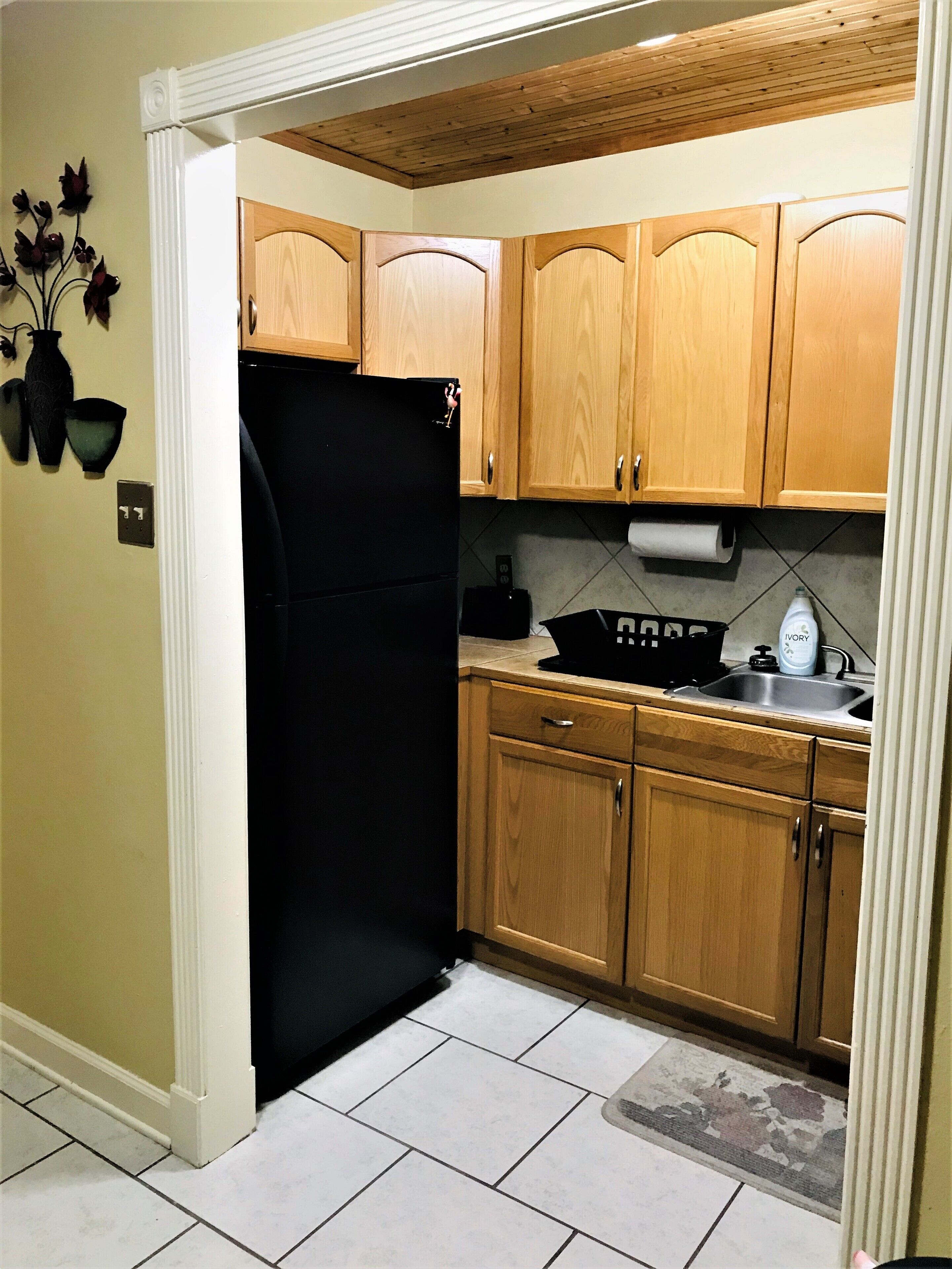 Una cocina con armarios de madera, un refrigerador negro, un lavamanos y una pequeña encimera con un tostador y una botella de detergente Ivory.