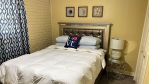 A neatly made bed with a white duvet and striped pillows, a tufted headboard, a bedside lamp, and a small decorative item on the nightstand. - 2 Blocks to LSU Gameday Shuttle. Best Downtown Location By Far (Baton Rouge)