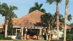 A building with a tiled roof, palm trees, and a water feature in the foreground. - Beautiful, modern home on golf course near South Padre Island (Laguna Vista)