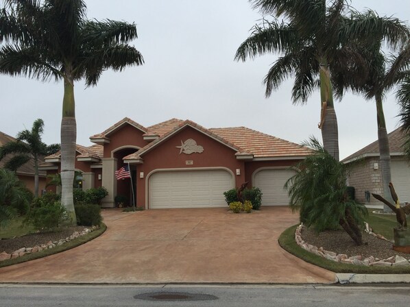 A residential house with a red facade, two garage doors, and a driveway leading to the entrance. - Beautiful, modern home on golf course near South Padre Island (Laguna Vista)