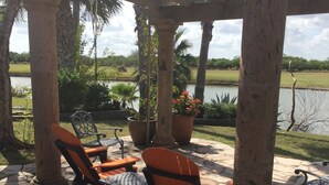 A covered patio area with stone flooring, two orange chairs, a table, and a view of a lake with palm trees and a grassy field in the background. - Beautiful, modern home on golf course near South Padre Island (Laguna Vista)