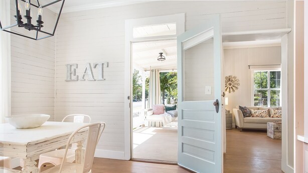 Amplia habitación con comedor que incluye una mesa y sillas blancas, una puerta abierta grande que conduce a una sala de estar con un sofá y un reloj de pared decorativo.