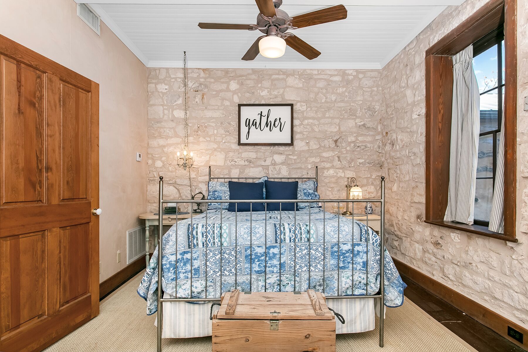 A bedroom with a stone wall, a bed with a metal frame, a wooden chest, a ceiling fan, and a window with curtains.