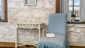 A room with a stone wall, a window with curtains, a chair with a cushion, and a small table with a book. - Itz Inn Back Courtyard Suite | Sleeps 2 | Next to Hondo's On Main Street (Fredericksburg)