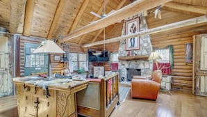 Interior rústico de una cabaña con techo de madera, chimenea de piedra, televisor y un sillón de cuero.