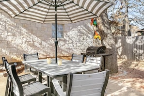 Une terrasse avec une table et des chaises, un parasol à rayures, un barbecue et un mur en pierre.