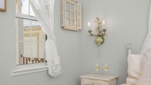 Un dormitorio con cama, una mesita de noche con dos copas de vino, un candelabro de pared con velas, y una ventana con cortina blanca.