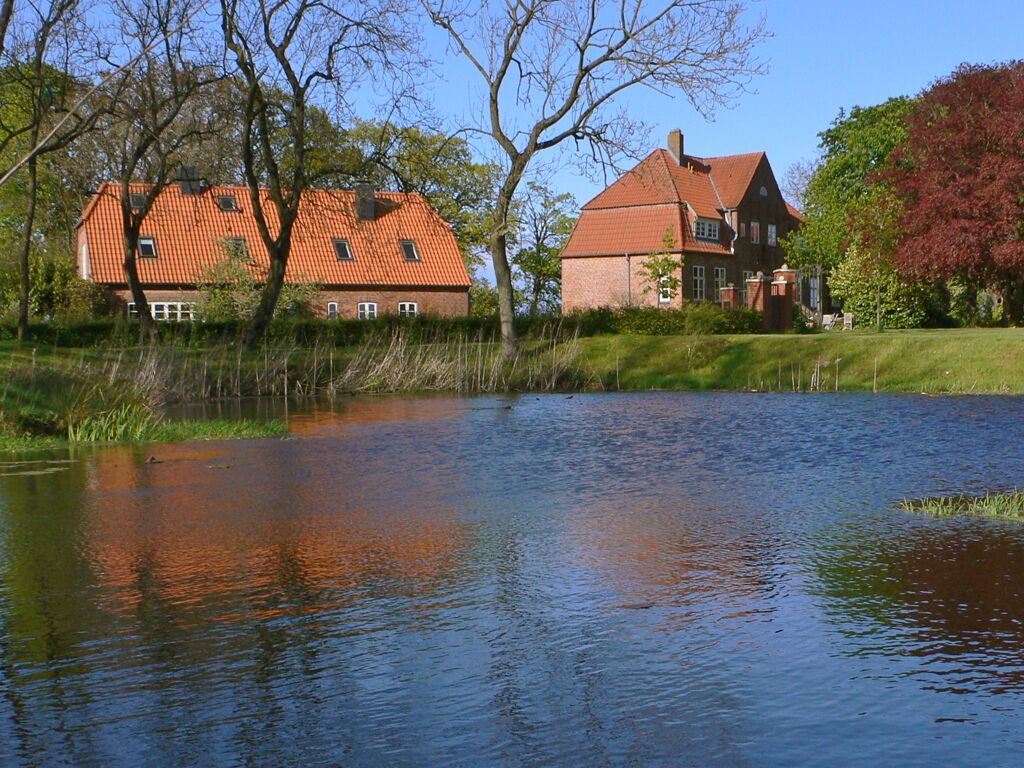 A house with a red roof and brick walls, situated next to a calm body of water with reflections.