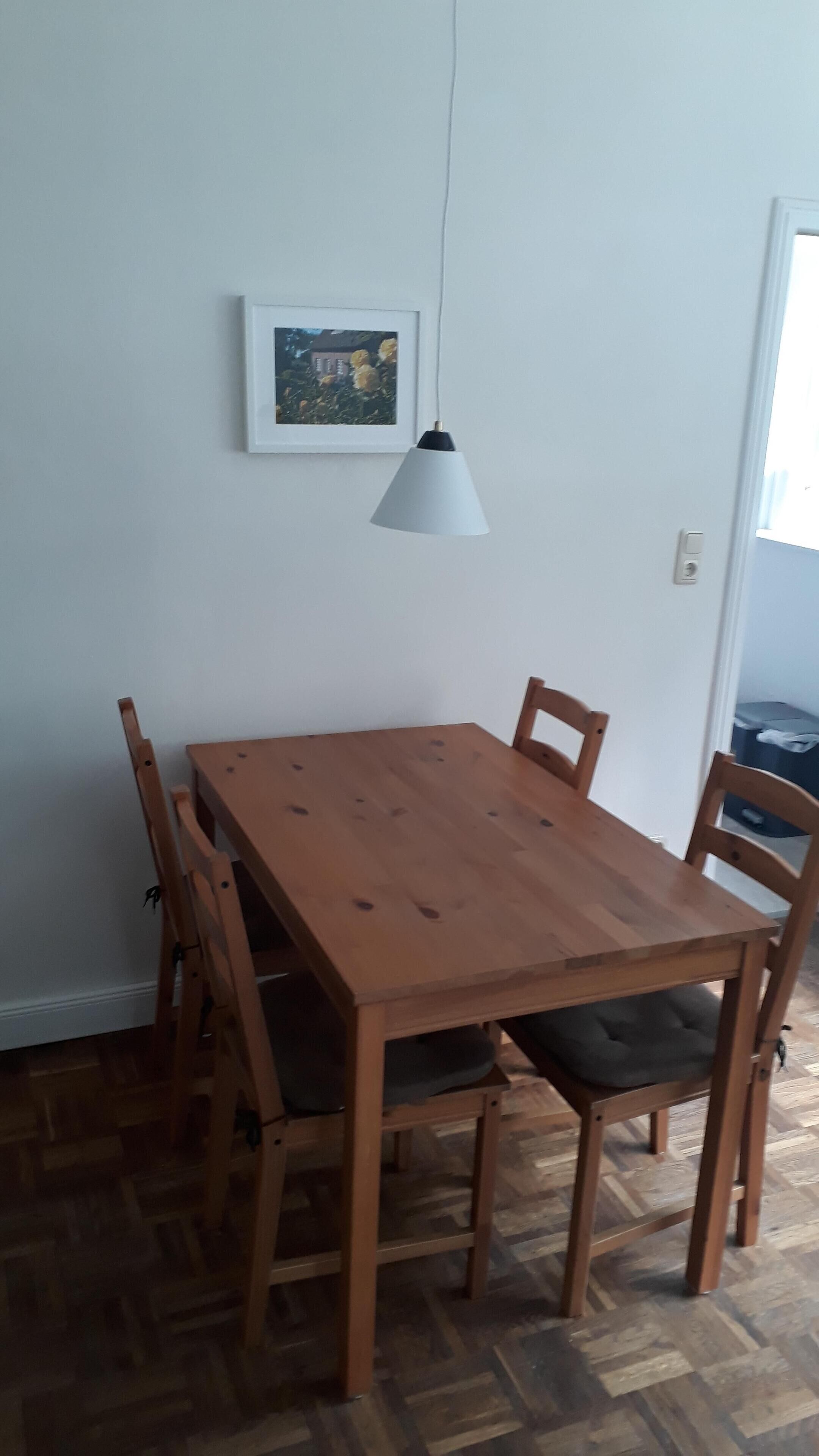 A wooden dining table with four chairs, a hanging light fixture, and a framed picture on the wall.