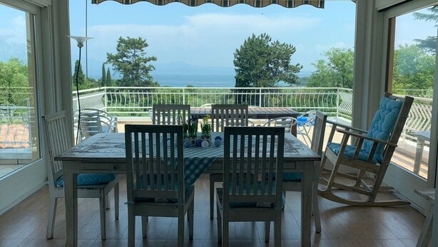 A covered outdoor dining area with a table and chairs, overlooking a scenic view of the sea and greenery.