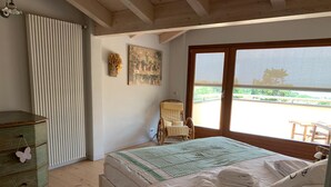 A bedroom with a bed, a chair, a radiator, a painting, and a view of the outside.