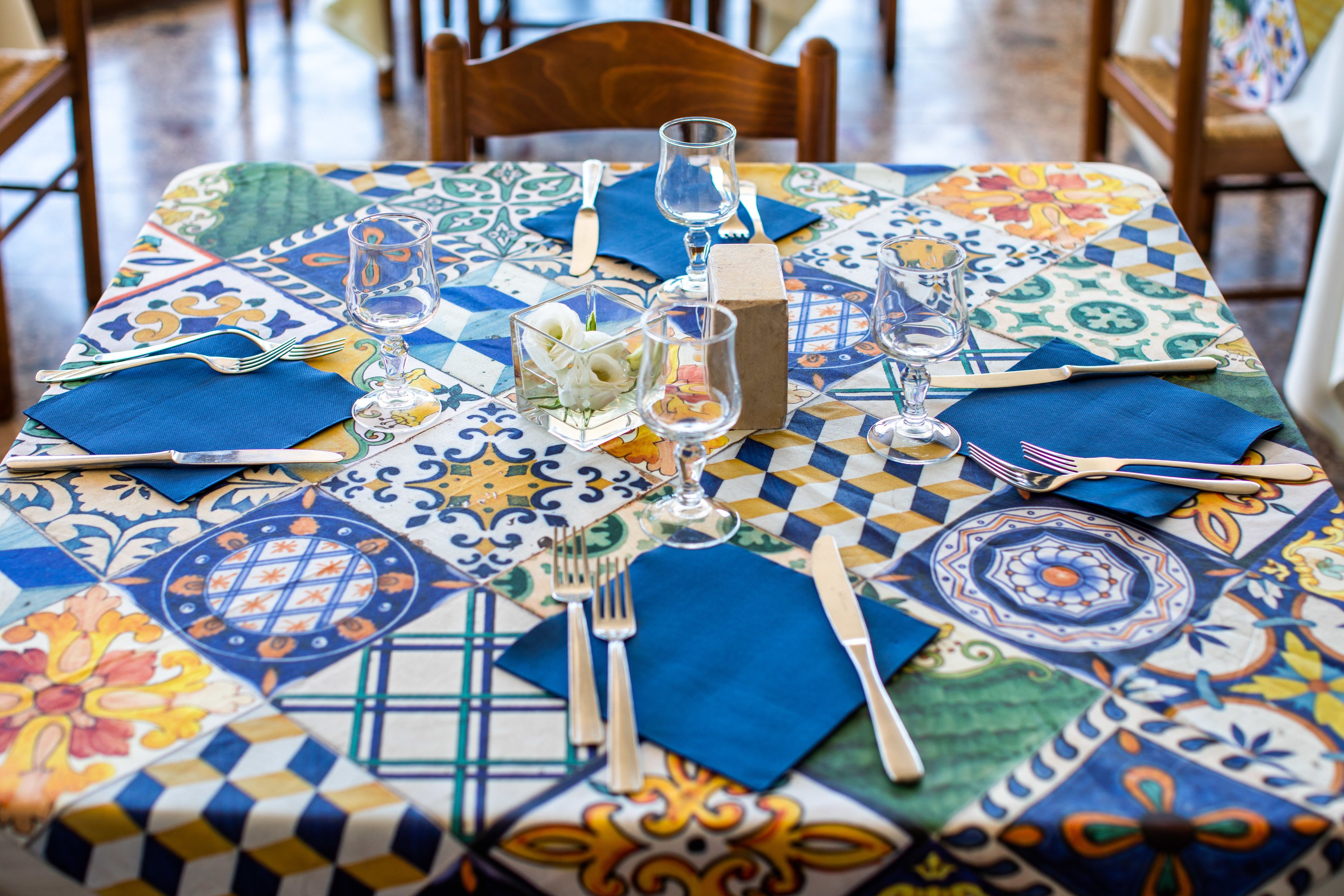 A table set with blue napkins, silverware, and clear glasses, covered by a colorful patterned tablecloth.