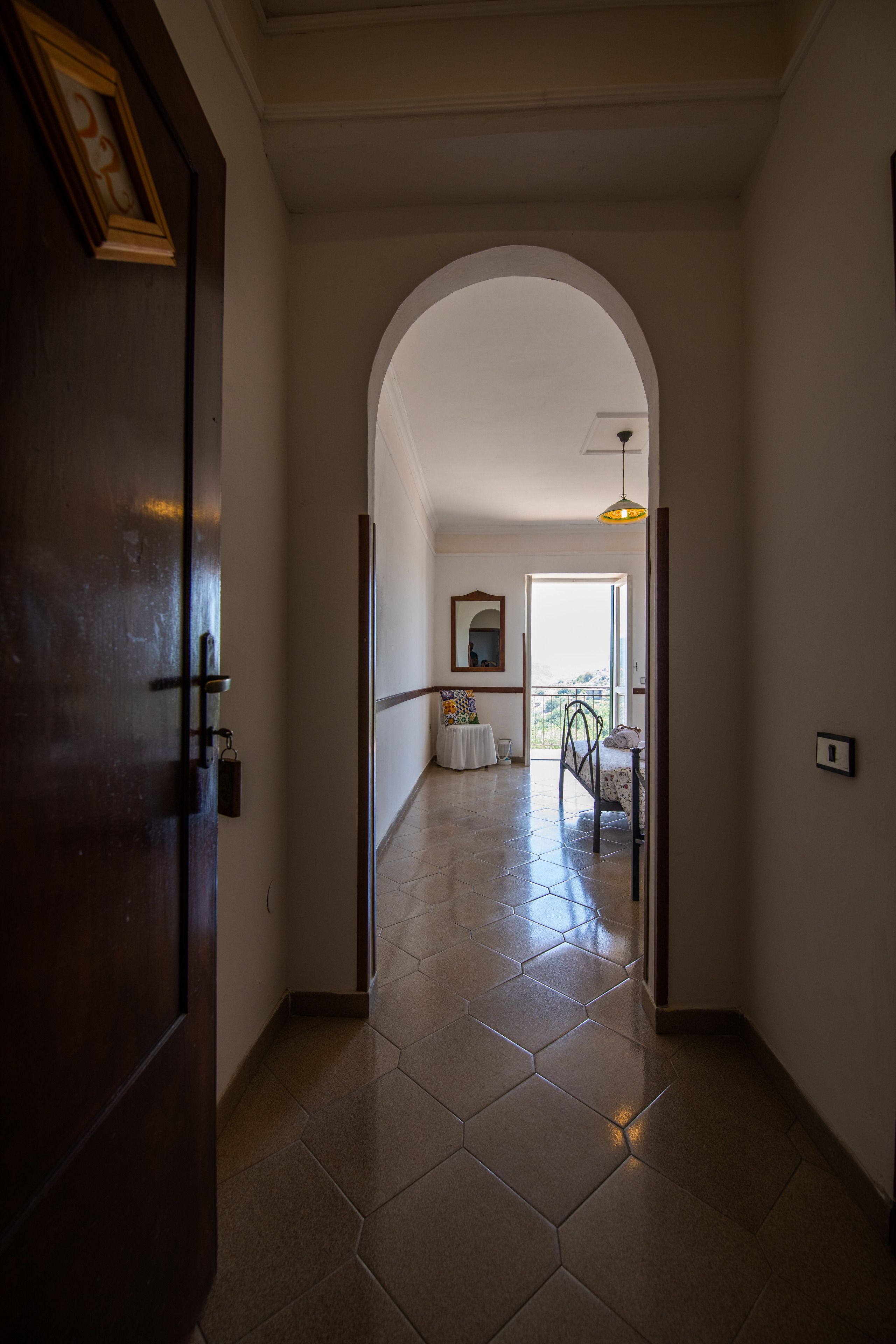 A hallway with a wooden door, a tiled floor, and an arched doorway leading to a balcony with a table and chairs.