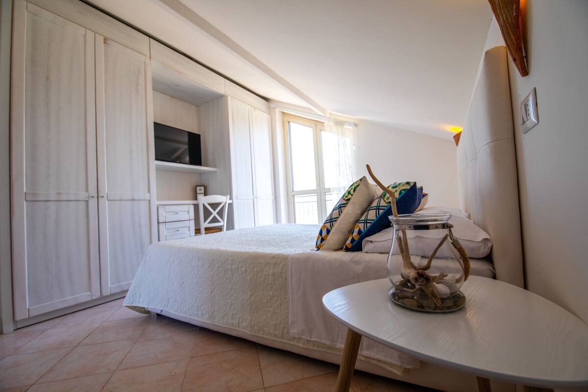 A bedroom with a bed, a small table with a decorative item, and a TV in the background.