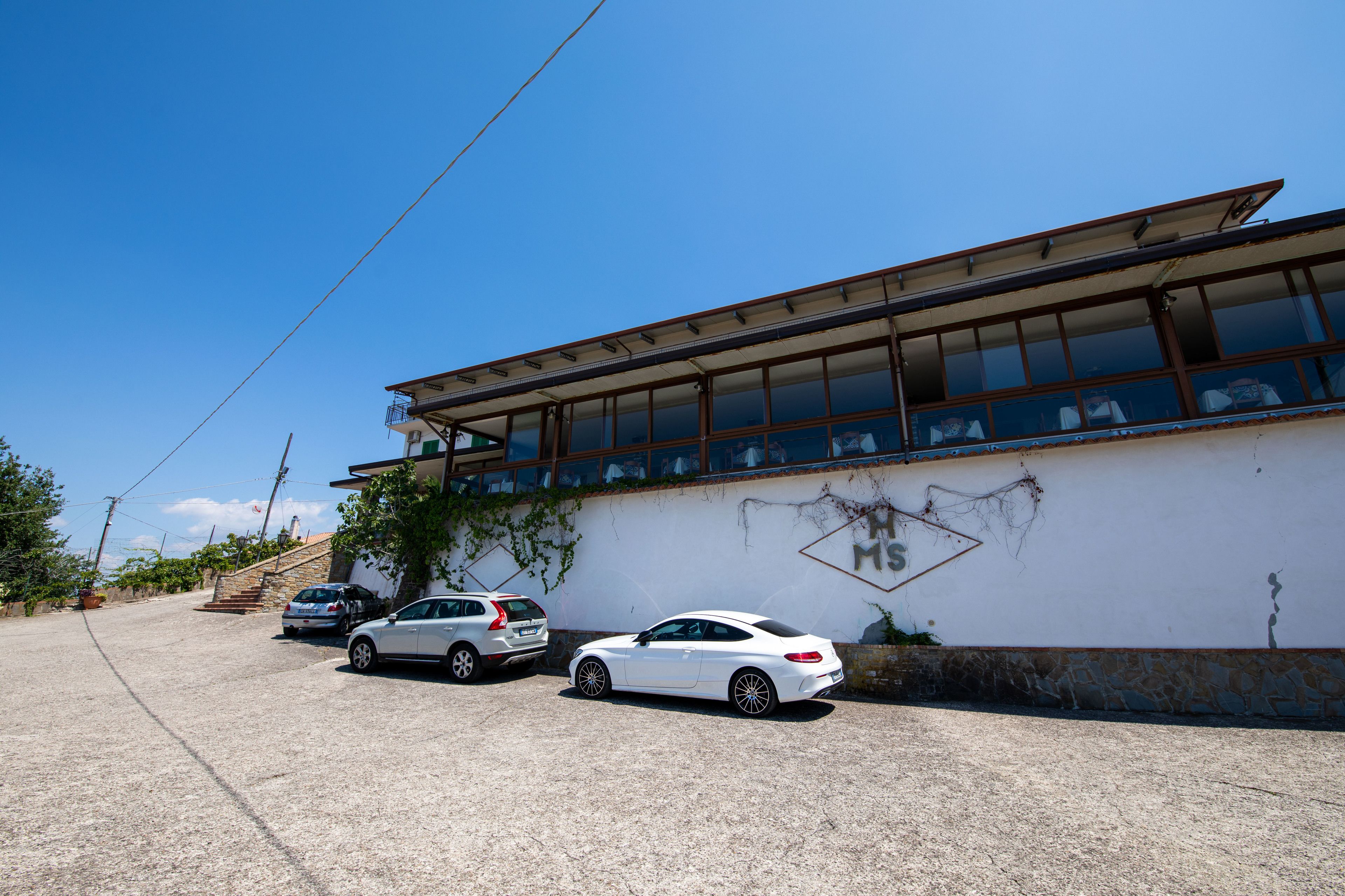 Un edificio con balcón, un coche blanco estacionado al frente y una pared de piedra con un letrero.