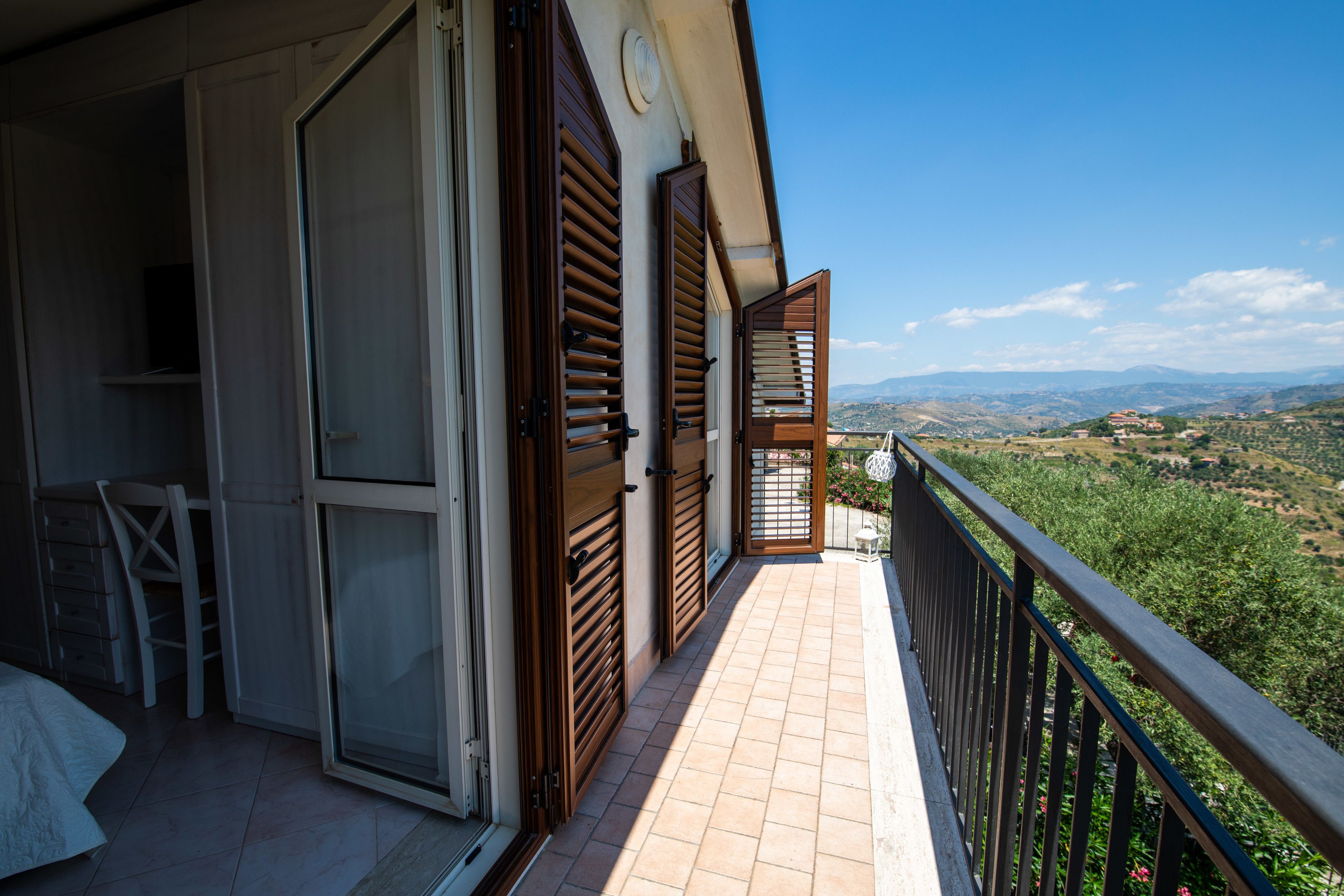 A balcony with wooden shutters, a tiled floor, and a view of a hilly landscape.