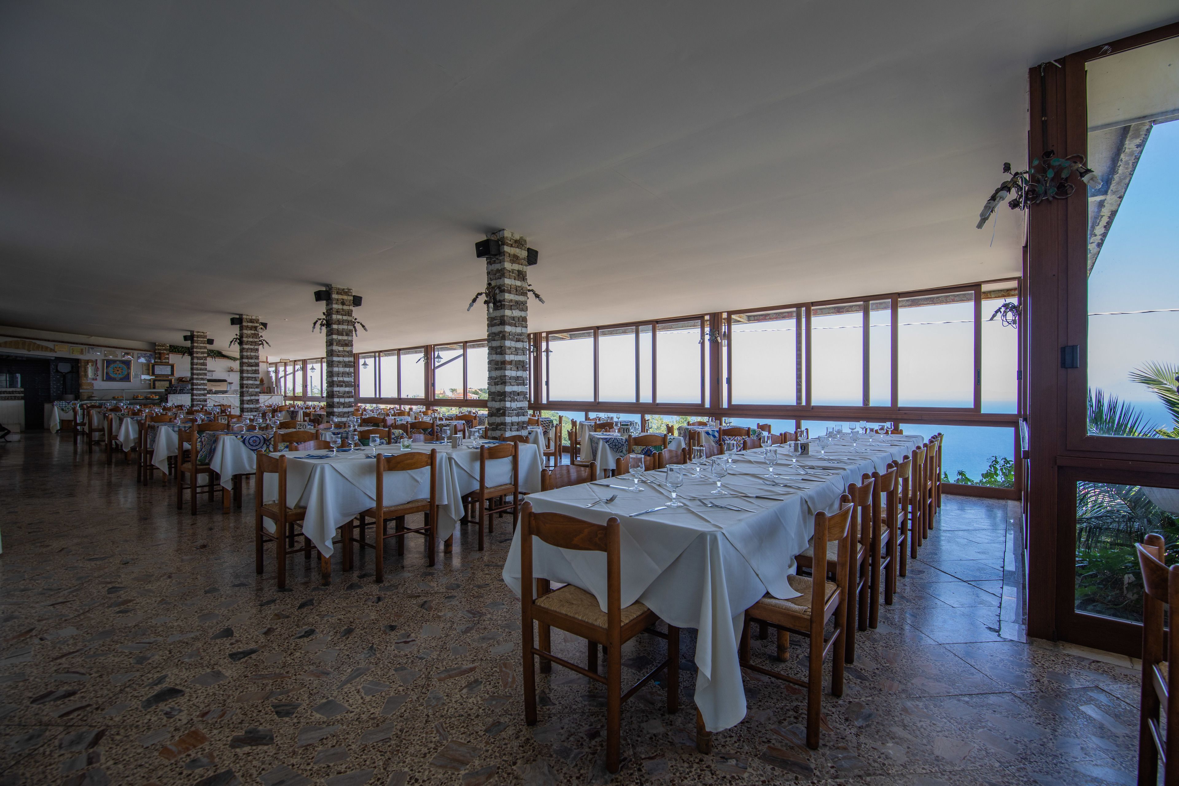 A spacious dining area with multiple tables set for dining, large windows offering a view, and a high ceiling.