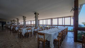 A spacious dining area with multiple tables set for dining, large windows offering a view, and a high ceiling.