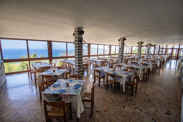 Un restaurant doté de nombreuses tables et chaises, de grandes fenêtres donnant sur l’océan et de piliers en pierre.
