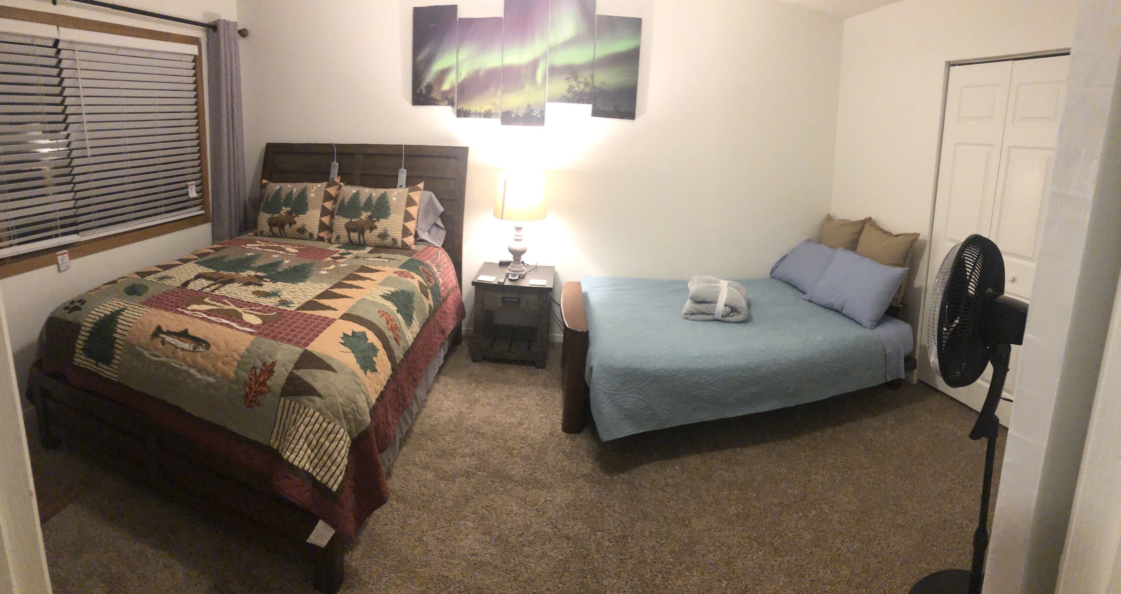 Een slaapkamer met een bed, nachtkastje, lamp, ventilator en een raam met jaloezieën.
