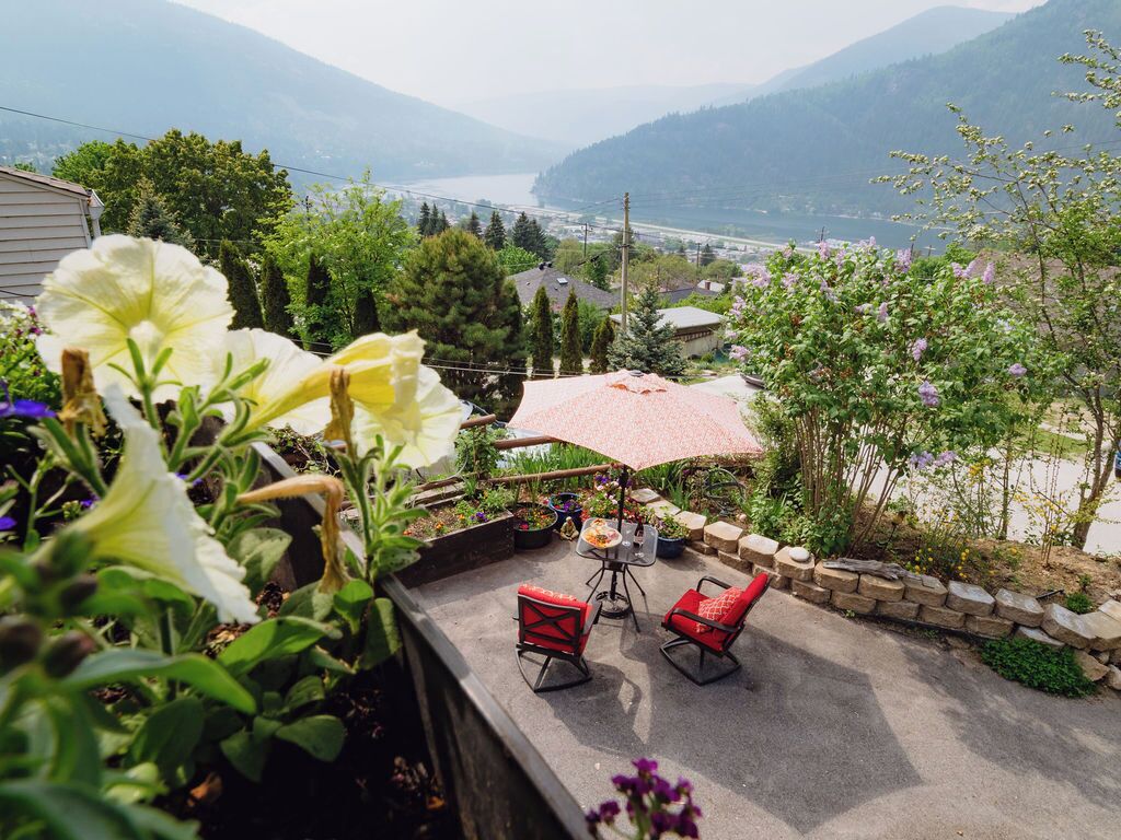 Une terrasse avec deux chaises rouges et une table, entourée de plantes et de fleurs en pot, d’où l’on peut admirer un paysage magnifique composé de montagnes et d’une étendue d’eau.