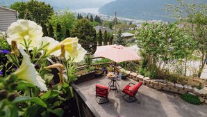 En terrasse med to røde stole og et bord, omgivet af krukker med planter og blomster, med udsigt over et naturskønt landskab med bjerge og et vandområde.