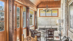Una habitación acogedora con pared de piedra, techo de madera y un ventanal grande. Hay un sofá, dos sillones de mimbre y una mesa con lámpara.