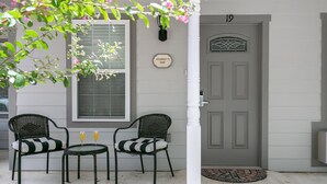 Une maison grise avec une porte blanche, deux chaises en osier, une petite table et une fenêtre donnant sur un beau paysage.