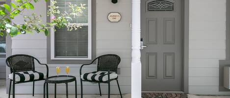 A gray house with a white door, two wicker chairs, a small table, and a window with a view.