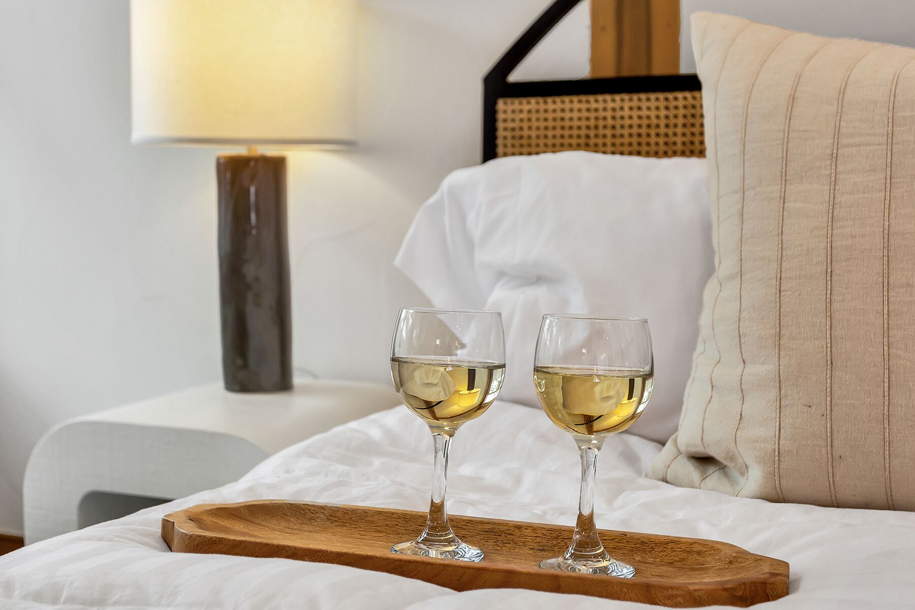 Dos copas de vino blanco sobre una bandeja de madera en una cama con sábanas blancas.