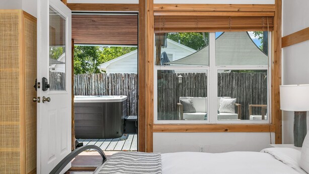 Un dormitorio con un ventanal grande, una puerta de madera, un jacuzzi y vistas a un patio trasero con valla y mobiliario exterior.