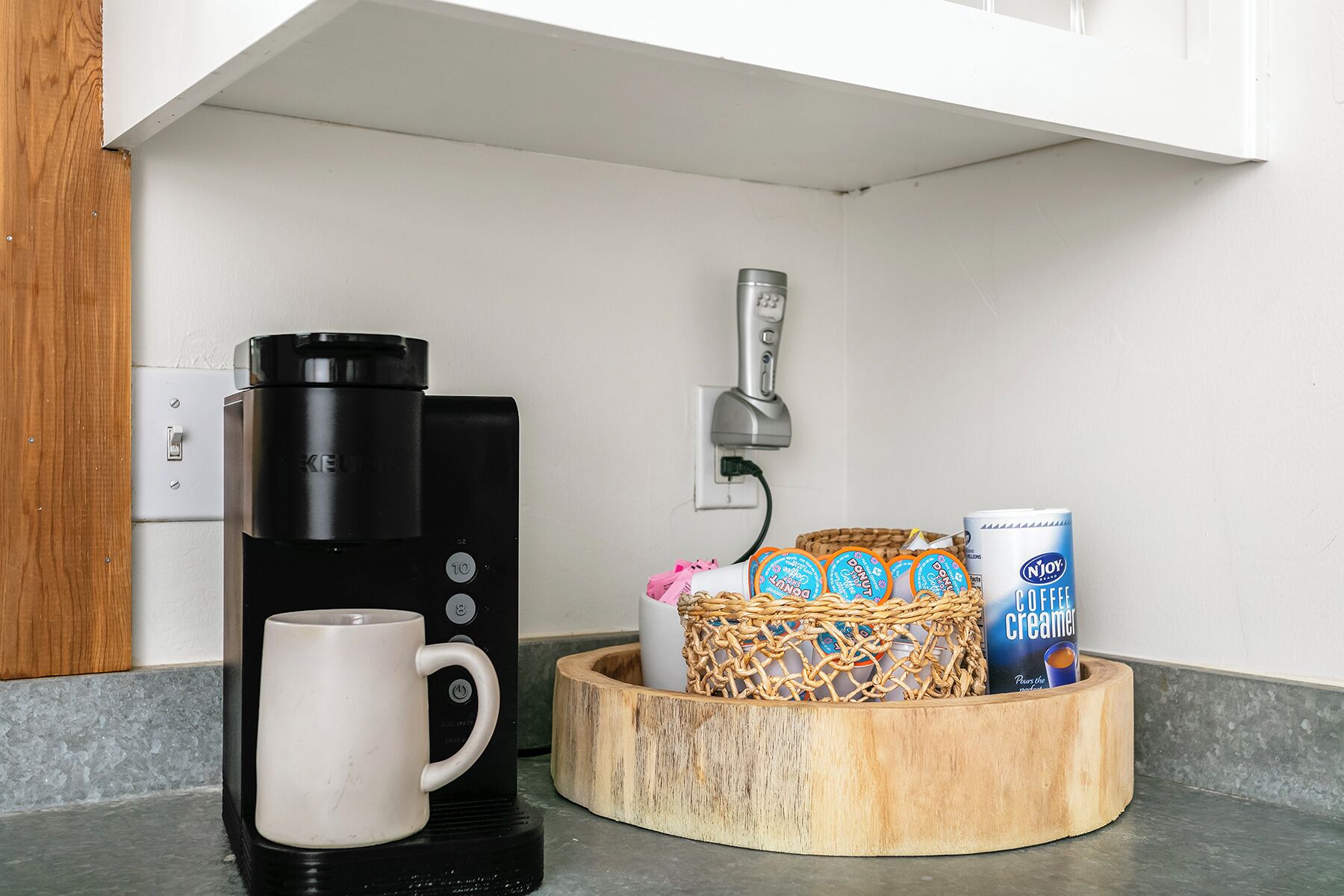 Una cafetera, una taza, y una cesta con leche condensada para café y donuts en la encimera de la cocina.