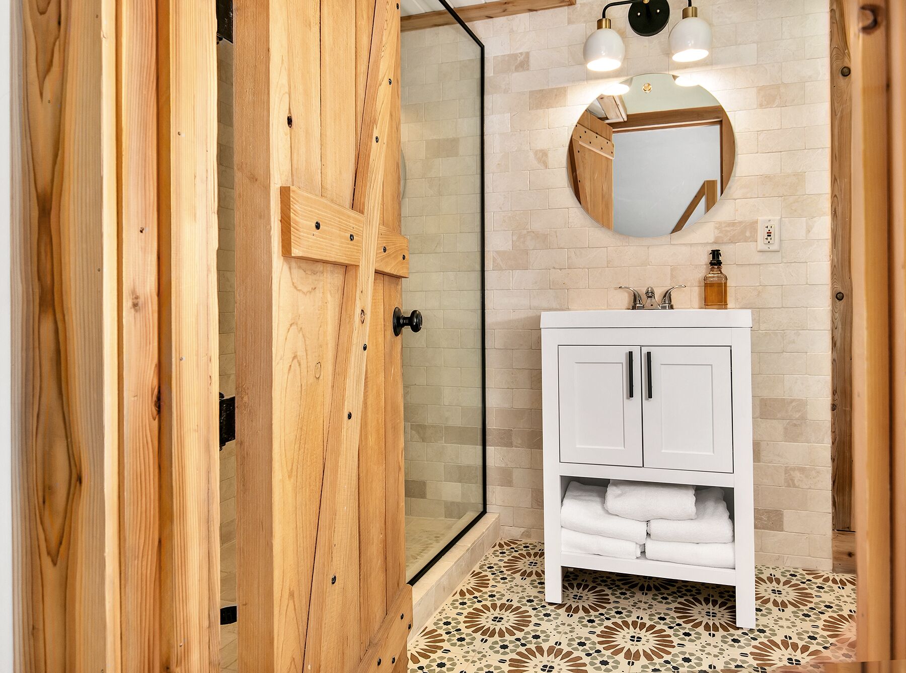 Un baño con puerta de madera, cabina de ducha con vidrio, tocador blanco con espejo y baldosas de suelo estampadas.