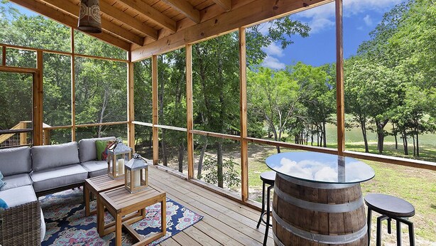Amplia terraza de madera con sofá, mesas y un barril, con vistas a un paisaje verde y exuberante.