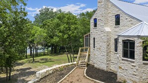 Un edificio de piedra con techo metálico, una escalera de madera que lleva a una puerta y vista a un lago rodeado de árboles.