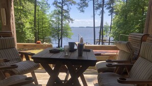 Une terrasse en bois avec une table et des chaises, donnant sur un lac bordé d’arbres et sur lequel on aperçoit un quai au loin.