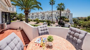 Een terras met een ronde tafel, twee grijze fauteuils met kussens, een bloempot en een fles champagne. Achtergrond: een wit gebouw met meerdere balkons en palmbomen.