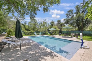 Una piscina rectangular con una terraza de baldosas, rodeada de árboles y un patio con mobiliario exterior.