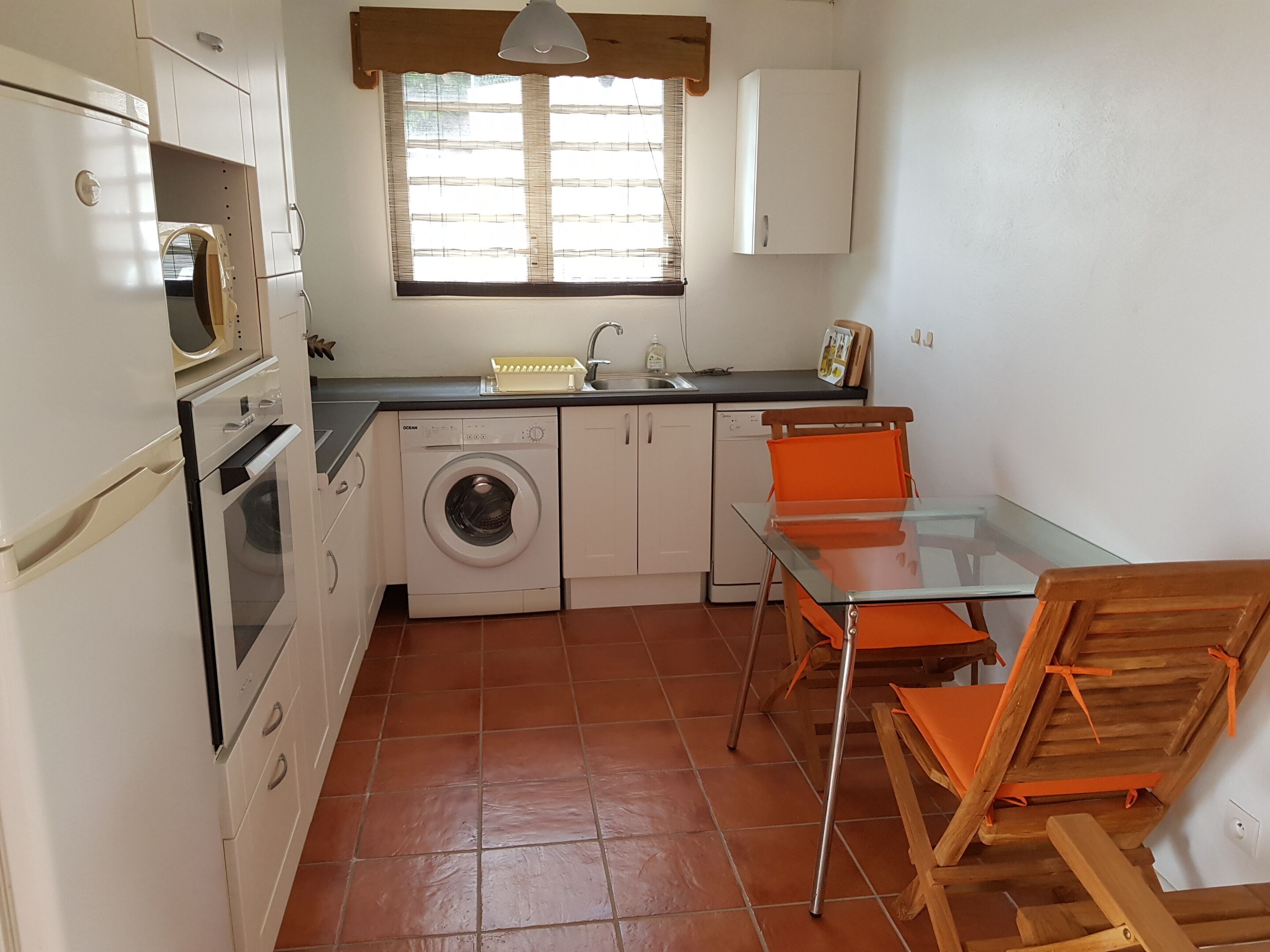 Una cocina compacta con lavadora, lavavajillas, una pequeña mesa de comedor con sillas y una ventana con persianas.
