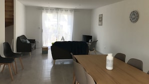 A modern living room with a wooden dining table, black and grey chairs, a black sofa, and a large window with sheer curtains. - New house with beautiful sea view (Groix)